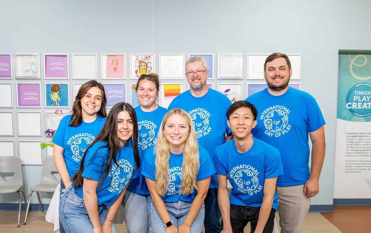 MAGNA-TILES team smiling in blue International Day of Play shirts in front of framed children's art
