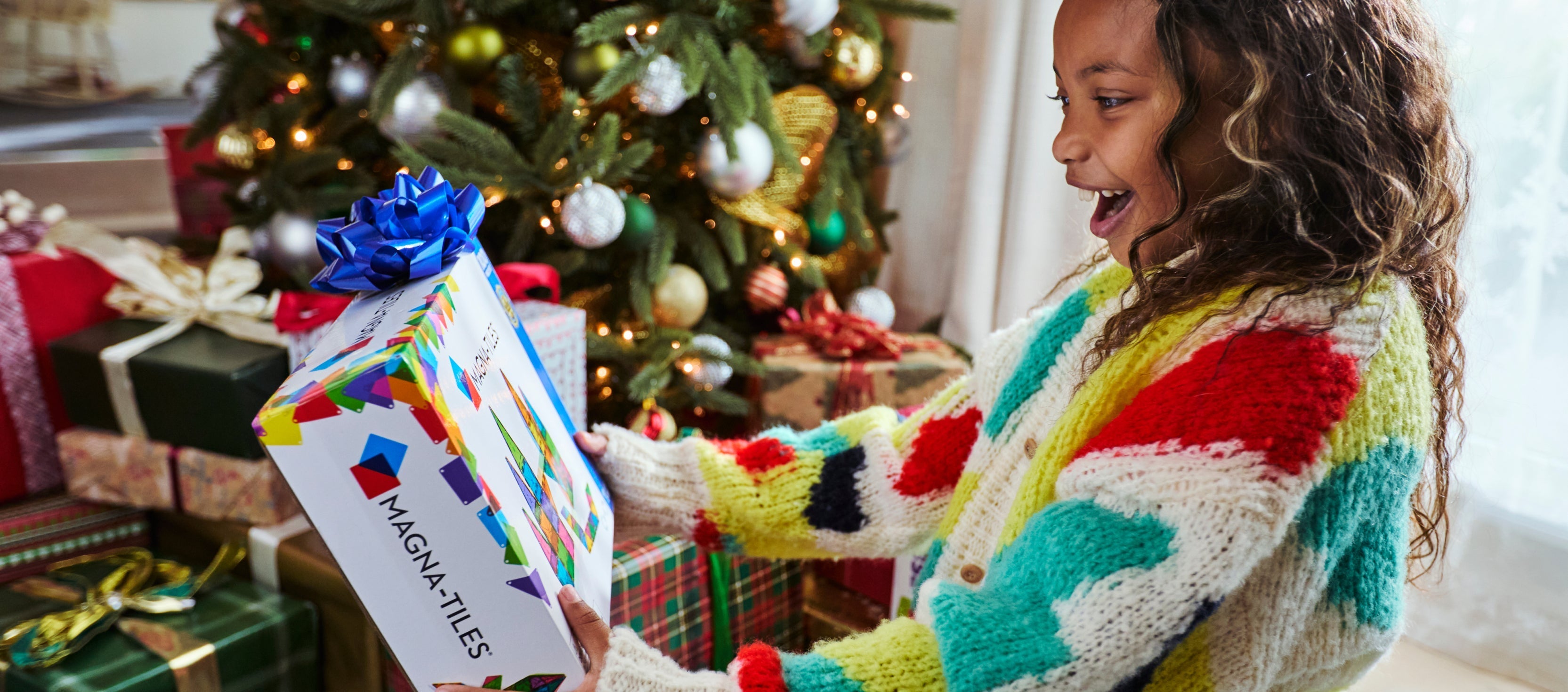 Girl excitedly holding a MAGNA-TILES gift box in front of a decorated Christmas tree and wrapped presents