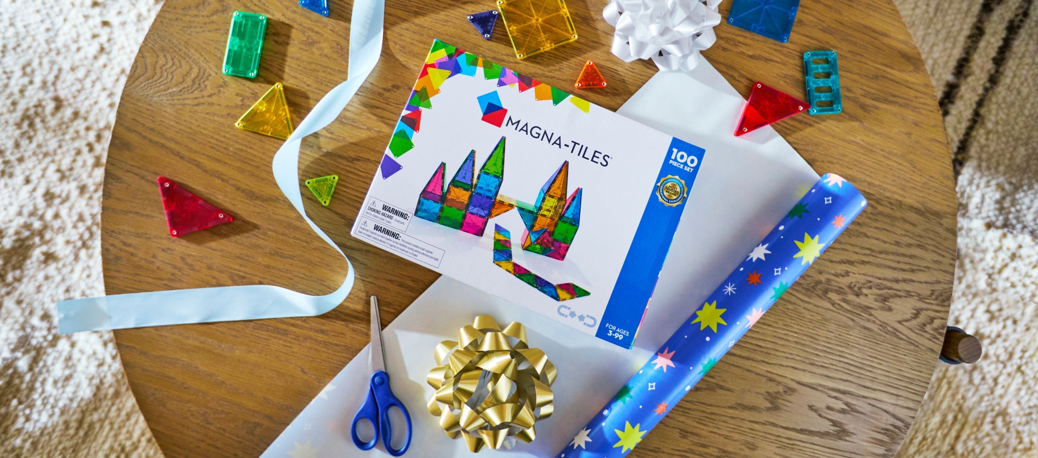 Angled overhead view of MAGNA-TILES 100-piece set box showing colorful translucent tiles and gift wrap on a wooden table
