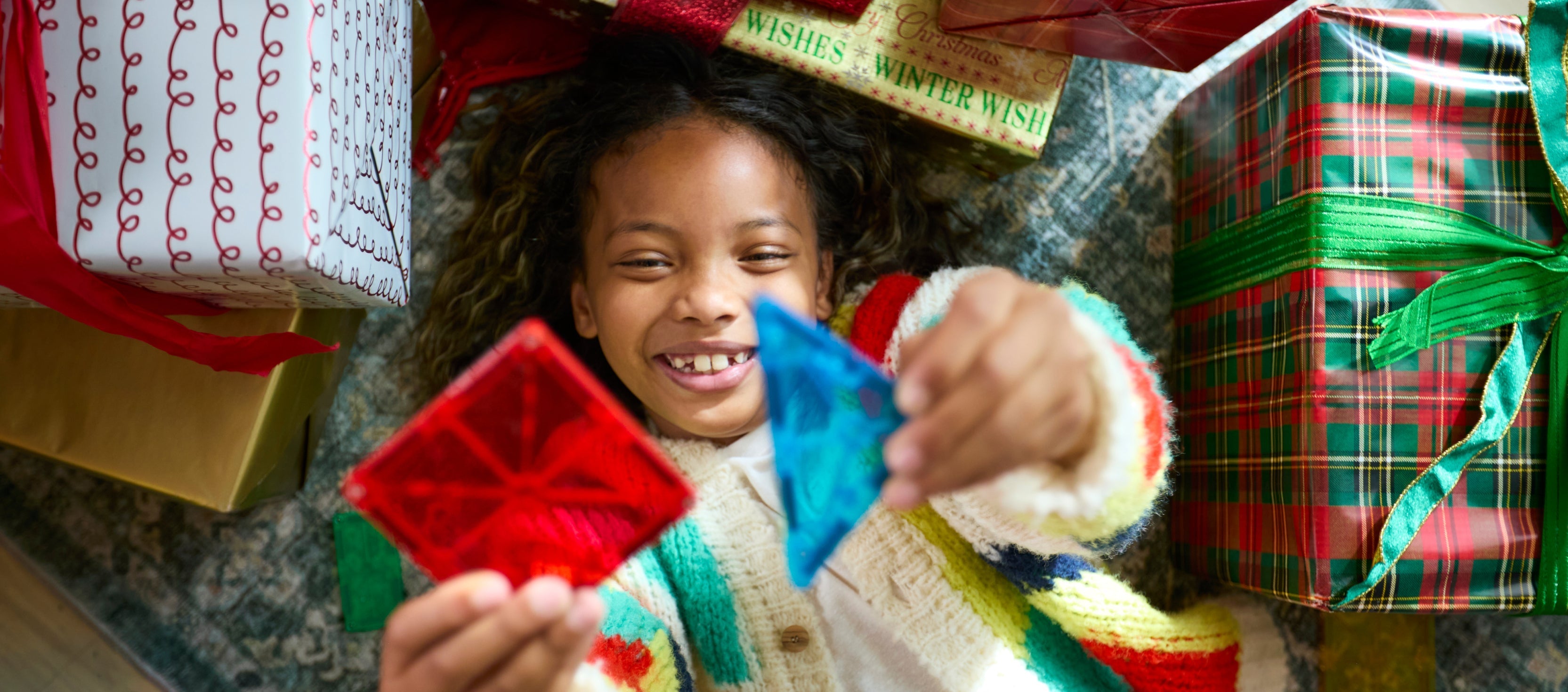 Smiling child lying among wrapped holiday gifts holding red and blue Magna-Tiles magnetic tiles