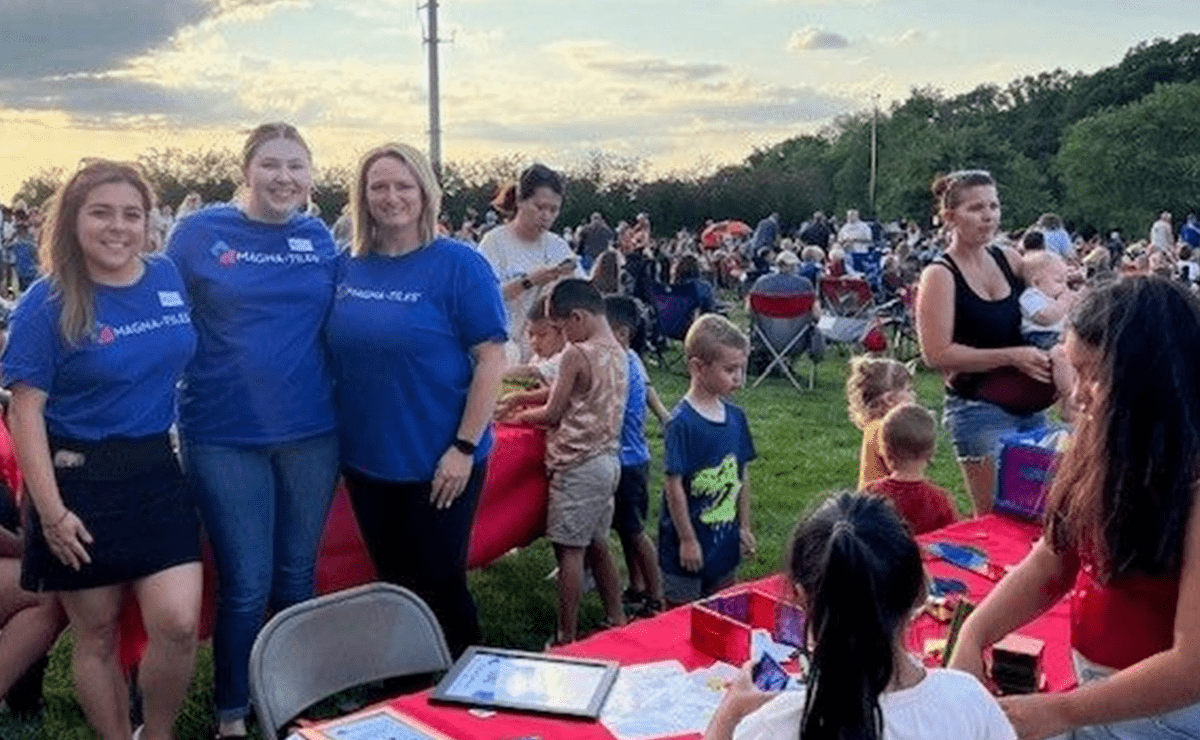 Three MAGNA-TILES staff standing by a play table while children play at an outdoor community event at sunset