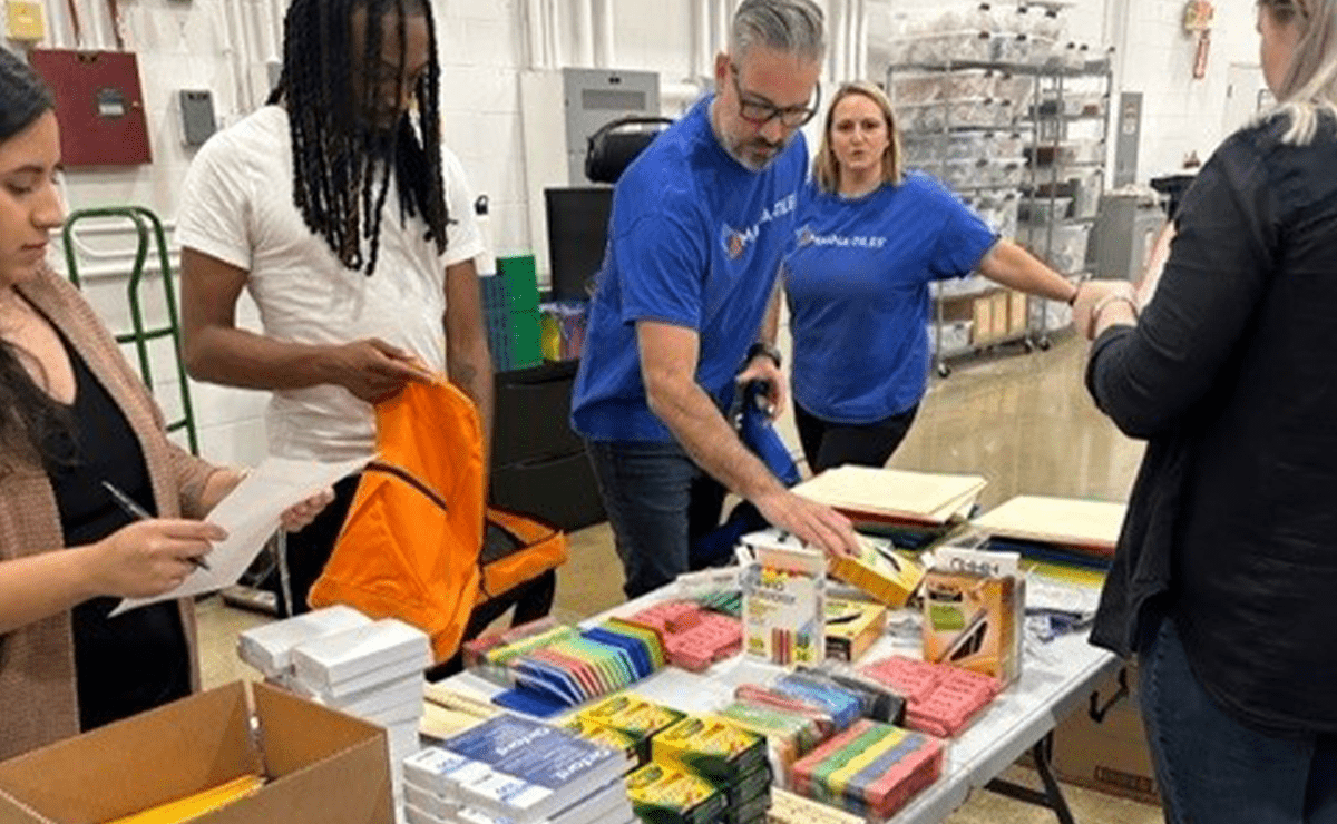 Volunteers packing school supplies into bright orange backpacks at a table in a warehouse