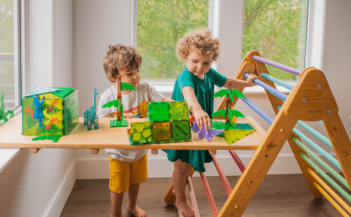 Two children play with green translucent magnetic tiles on a wooden plank across a colorful climbing frame by a window