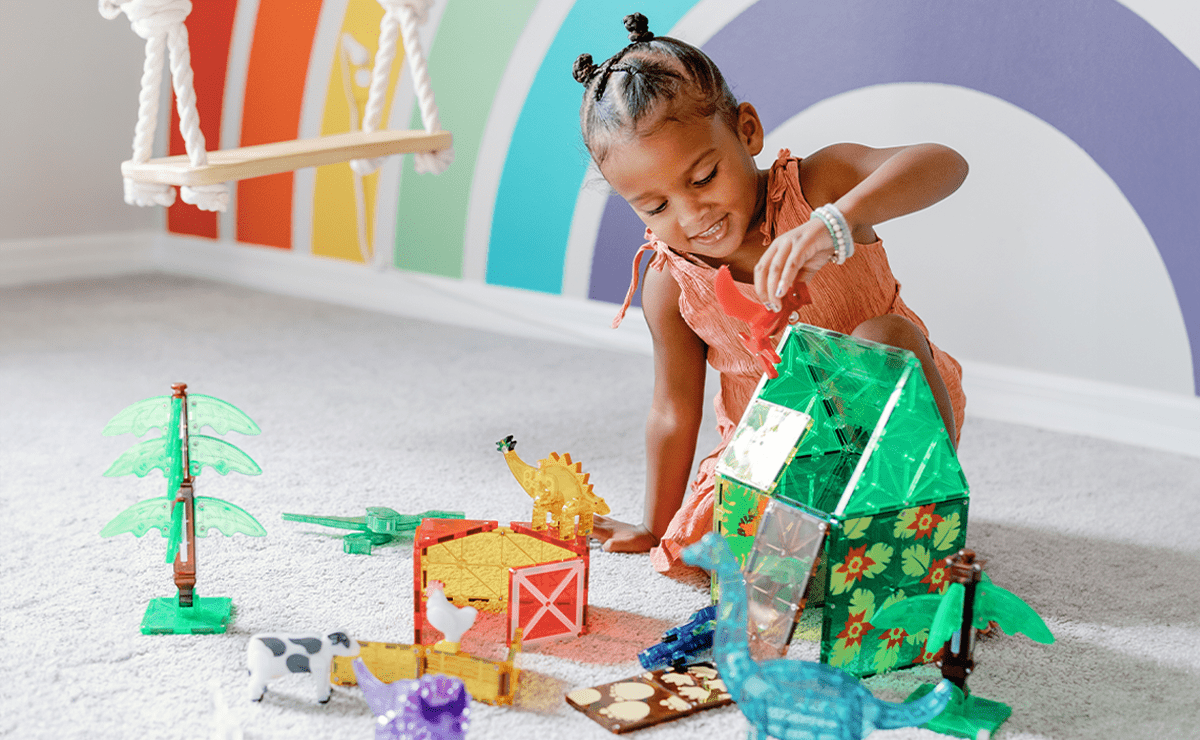 Young girl playing with a green MAGNA-TILES house and animal figures on a carpeted floor in a colorful playroom