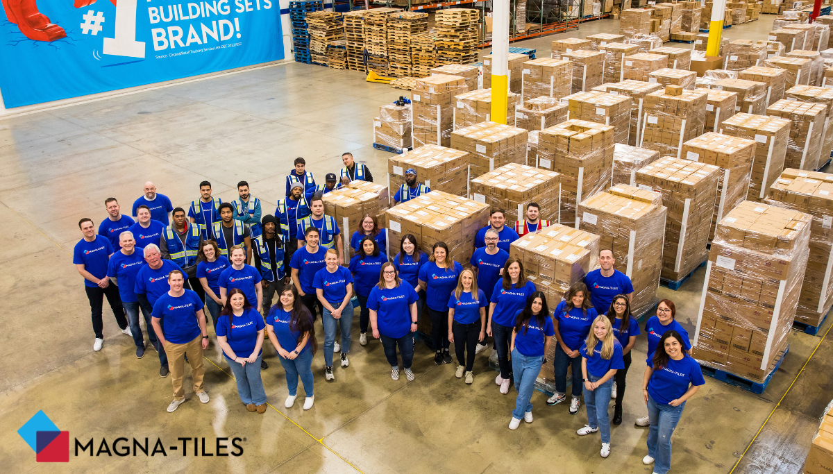 MAGNA-TILES employees in blue shirts pose in a warehouse among stacked pallets of boxed sets