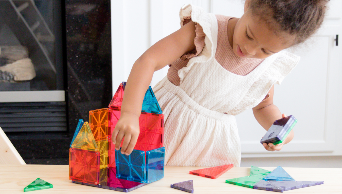 Girl assembling a colorful tower of translucent MAGNA-TILES on a wooden table in a bright kitchen