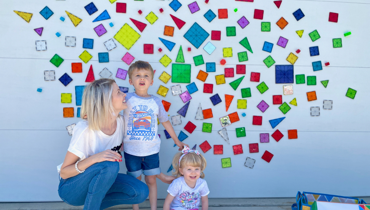 Woman kneeling with two young children in front of a white wall covered with scattered colorful MAGNA-TILES shapes