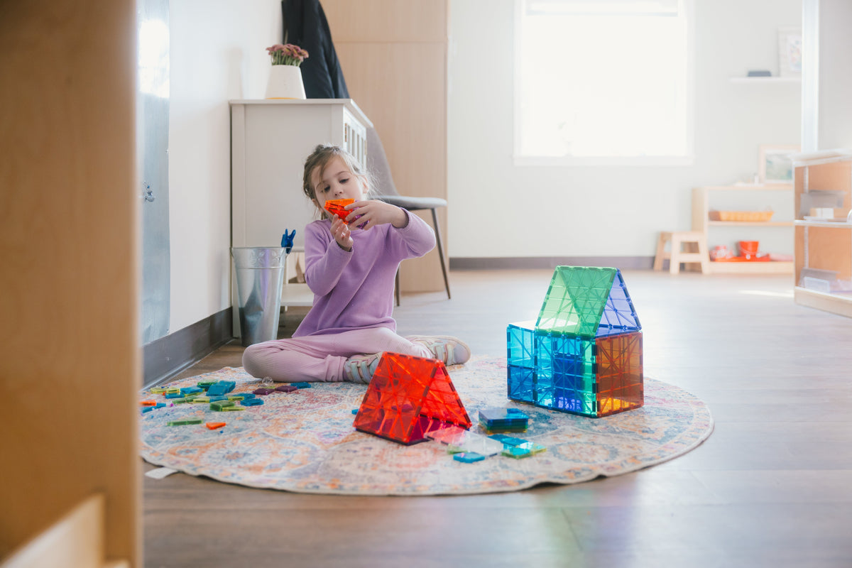 Girl sitting cross‑legged on a rug assembling red and blue translucent Magna‑Tiles in a bright Montessori classroom