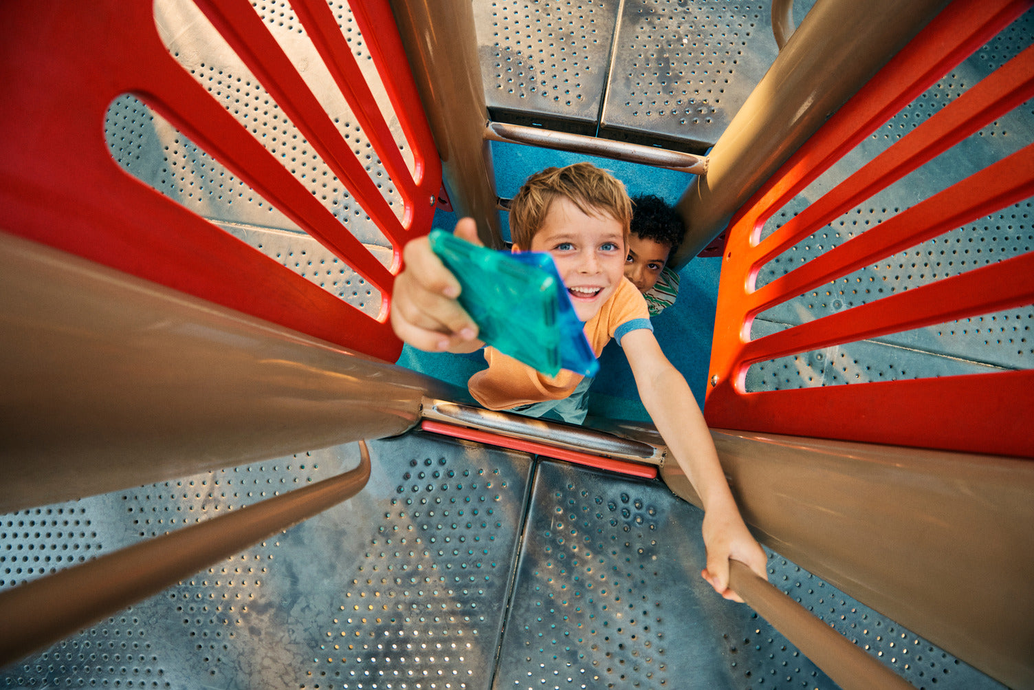 Two boys inside a playground tower, one smiling and reaching up while holding a translucent blue Magna-TILES triangle