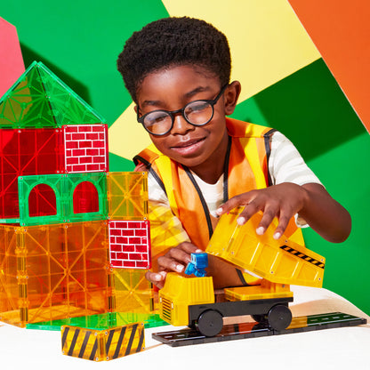 Child in orange vest plays with MAGNA-TILES Builder XL, loading a yellow dump truck next to a translucent tile structure