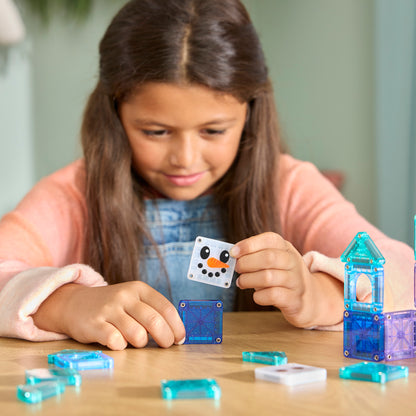 Girl playing with microMAGS Winter Wonder tiles on a wooden table, holding a snowman tile above blue translucent squares