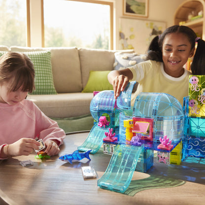 Two children playing with a translucent blue Undersea Adventure Magna-Tiles set on a living room table, showing domes, slides, a shark figure, and small character pieces