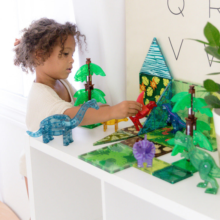 Child playing with MAGNA-TILES dinosaur set on a white shelf, arranging translucent dinosaur figures and green palm-tree tiles