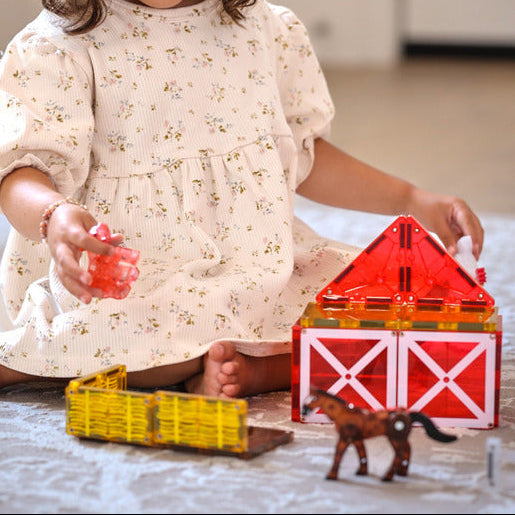 Child playing with MAGNA-TILES farm set, building a red barn with a yellow magnetic fence and a brown horse on a rug