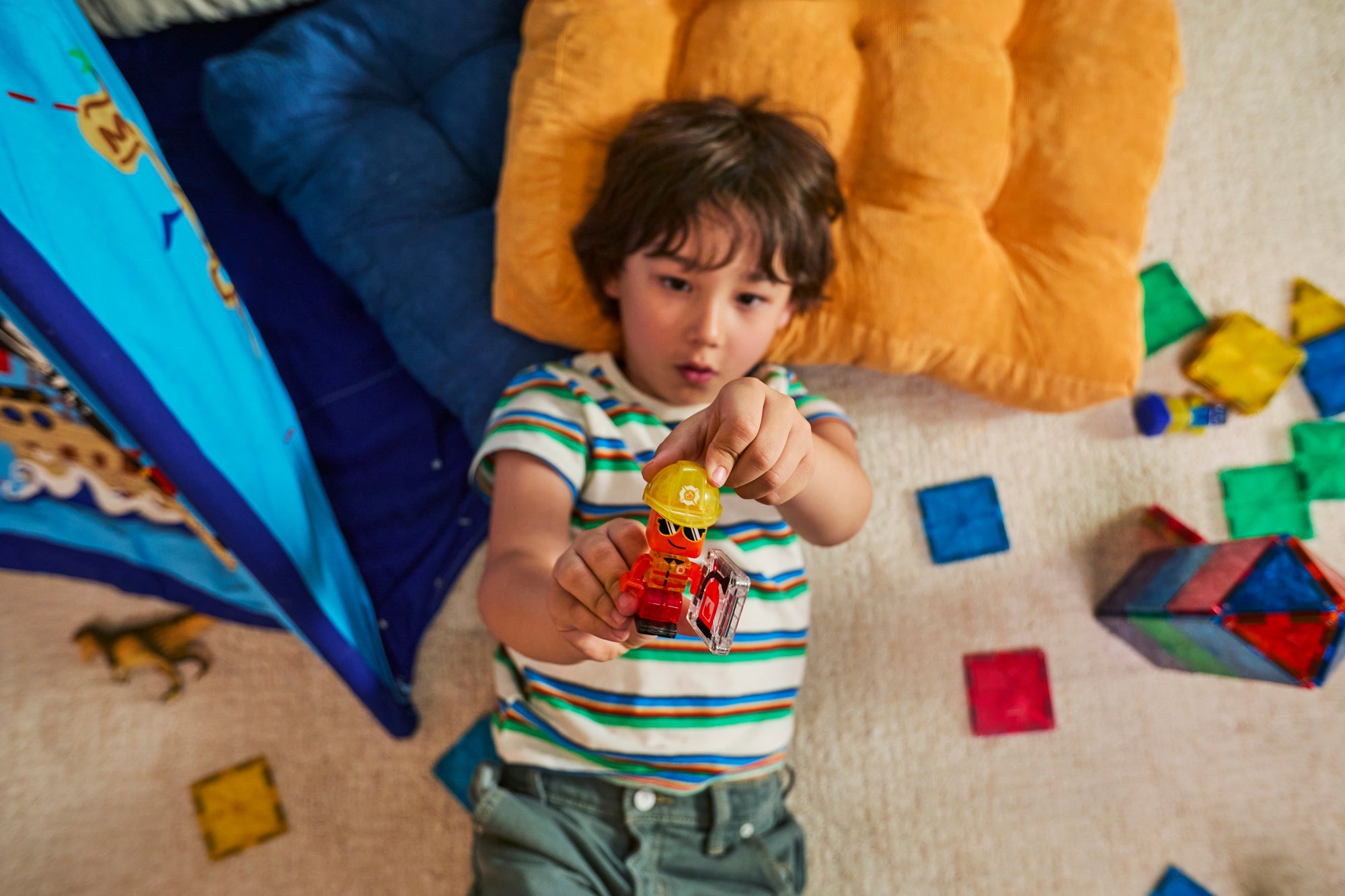 Young boy lying on a carpet reaching forward to show a toy figure, surrounded by colorful magnetic tiles and pillows
