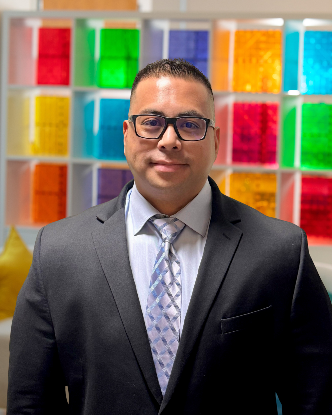 Man in suit and patterned tie smiling in front of colorful translucent tile wall in office