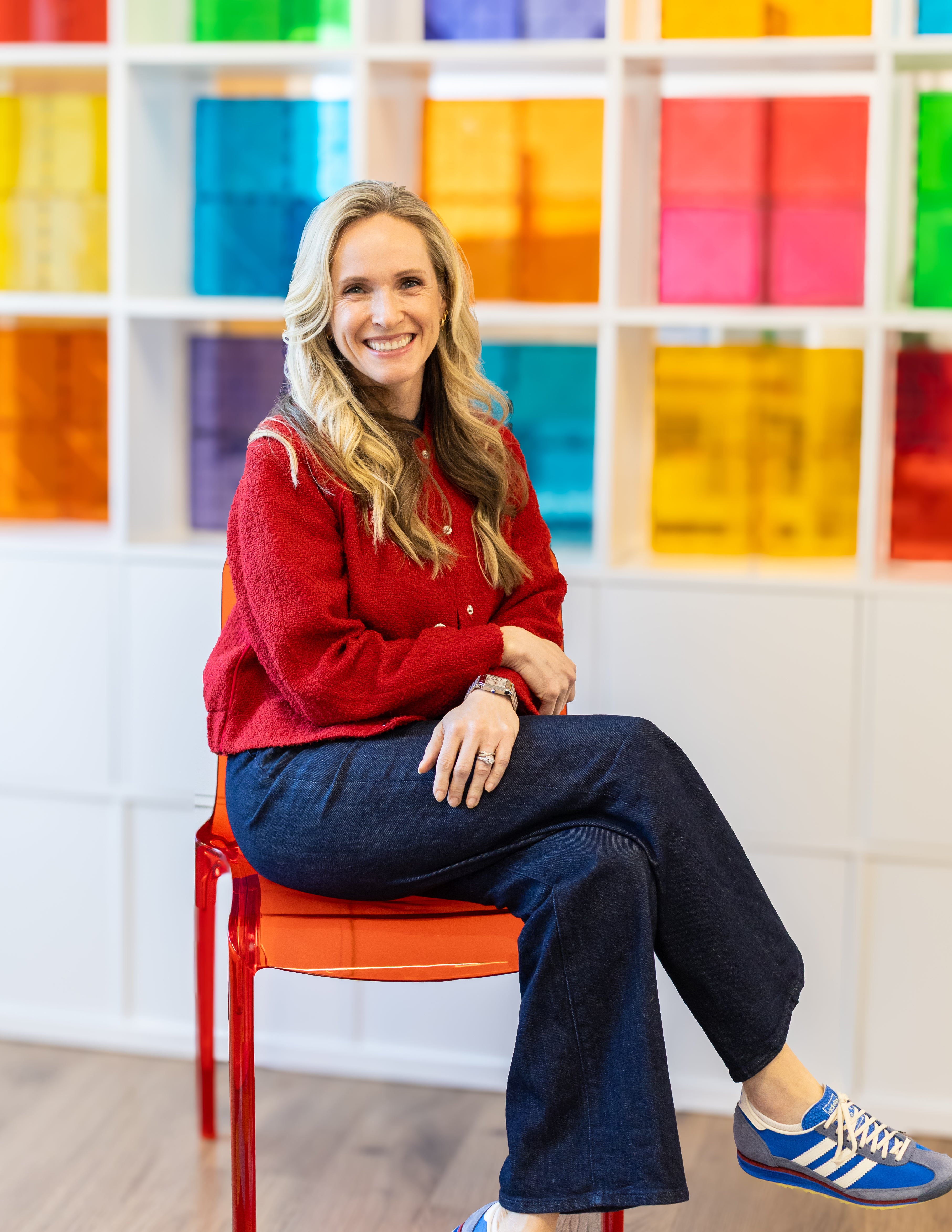 Smiling woman sitting with legs crossed on an orange chair in front of a wall of colorful translucent tiles